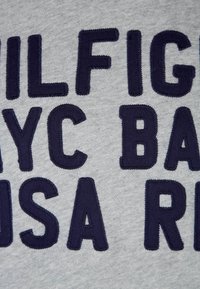 Šedá bavlněná mikina s výrazným vyšívaným textem v námořnicky modré barvě. Text zahrnuje "TOMMY HILFIGER NYC BASE USA RED" ve blokovém písmu.