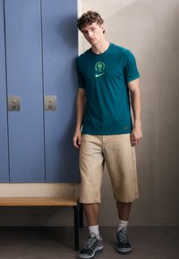 Teal athletic t-shirt with green logo and stripes, paired with light beige denim shorts, complemented by gray sneakers and white socks.