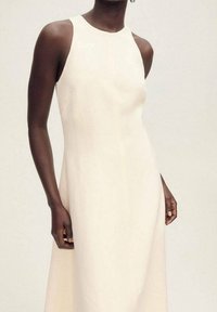 Kermanvärinen ilmaisin mekko, jossa on korkea kaula-aukko, sileä pinta, virtaava muotoilu ja hienovarainen A-linja. Ei näkyviä kuvioita tai koristeluja.