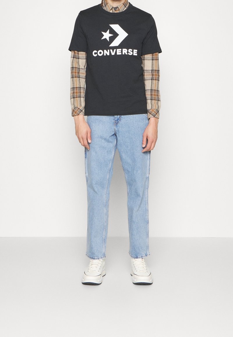 Homme portant un t-shirt Converse noir par-dessus une chemise à carreaux, un jean bleu clair et des baskets blanches, debout devant un fond blanc.