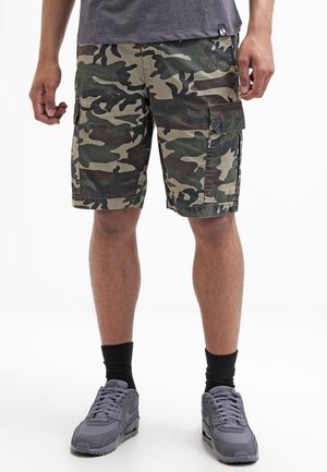 Homme portant un short cargo camouflage vert et marron, des chaussettes noires et des chaussures de sport grises, debout sur un fond blanc.