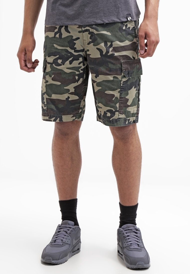Homme portant un short cargo camouflage vert et marron, des chaussettes noires et des chaussures de sport grises, debout sur un fond blanc.
