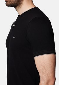 Czarna koszulka polo z krótkim rękawem, z zapięciem na guziki, teksturowana tkanina i kontrastowe przeszycia na rękawach. Logo na klatce piersiowej.