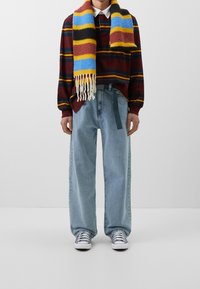 Gestreiftes, kastanienbraunes Langarmshirt, blau-gelb gemusterter Schal, hellblaue weit geschnittene Jeans, graue Sneaker mit weißen Sohlen.