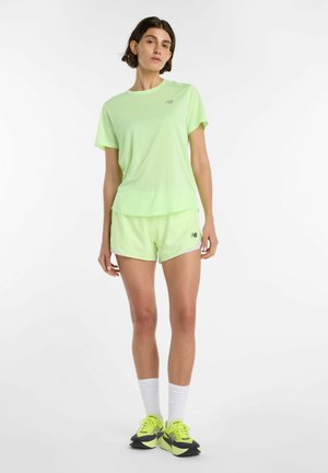 Donna in piedi che indossa una maglietta verde neon e pantaloncini New Balance, con calzini bianchi e scarpe da corsa neon su sfondo bianco.