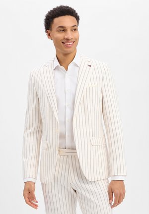 Junger Mann lächelnd, trägt einen weißen Anzug mit dünnen schwarzen vertikalen Streifen und ein weißes Hemd vor einem einfarbigen Hintergrund.