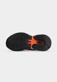 Semelle de chaussure de sport noire et orange avec motifs de relief texturés et clous en caoutchouc circulaires pour adhérence sur un fond gris clair uni.