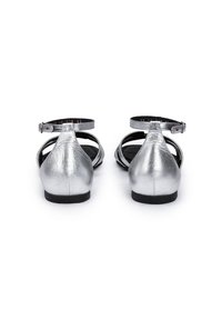 Paire de sandales plates métalliques argentées avec brides à la cheville, vues de dos sur un fond blanc.