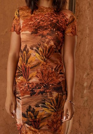 Vrouw in een getailleerde jurk met bruin, oranje en geel blad- en boompatroon, staand tegen een getextureerde bruine achtergrond.