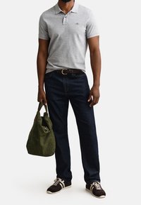 Homme portant un polo gris à manches courtes, jean bleu foncé, baskets marron, tenant un sac cabas en daim vert avec détail anneau.