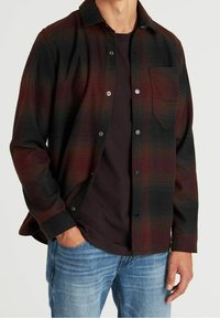 Camicia di flanella con motivo a quadri nero e bordeaux, chiusura con bottoni, taschino singolo sul petto, indossata sopra una maglietta scura, abbinata a jeans blu.