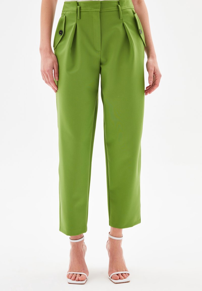 Pantalon vert taille haute à jambes droites avec plis et poches à rabat avant, porté avec des sandales blanches à talons hauts et lanières.