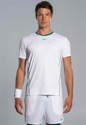 Uomo in piedi che indossa una maglia sportiva bianca e pantaloncini con rifiniture verdi, e un braccialetto bianco al polso sinistro.