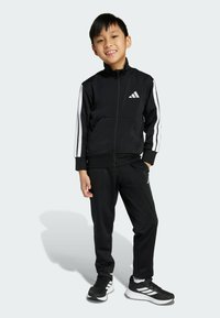 Černý sportovní teplákový komplet obsahující bundu na zip s bílými pruhy a logem, spárovaný s odpovídajícími kalhotami a černými sportovními botami.