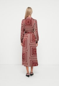 Maxi-mekko viininpunaisena, jossa on geometristen kuvioiden vaalean kermanväristä. Sitä koristavat pitkät hihat, vyöllinen vyötärö ja virtaava siluetti.