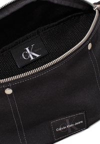 Melnas kanvas maisiņš ar rāvējslēdzēja aizdari. Iekšpusei ir acs dizains. Iekļauj logo etiketes, metāla detaļas un kontrastējošu baltu dūrienu.