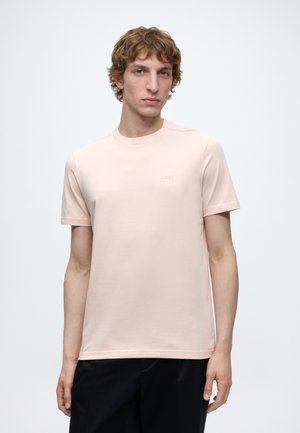 Jeune homme aux cheveux châtain clair bouclés, portant un t-shirt rose pâle à manches courtes et un pantalon noir, debout devant un fond blanc uni.