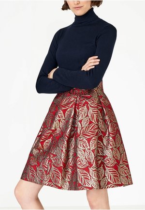 Jupe rouge avec un motif de feuilles en or et en argent, dotée d'une taille ajustée et d'un design évasé. Associée à un pull à col roulé noir.