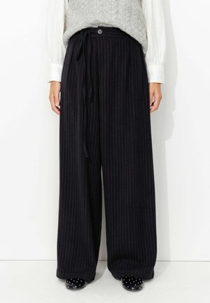 Pantalons à jambes larges, rayés bleu marine, avec une taille haute, une ceinture en tissu et une fermeture à un bouton. Associés à un gilet en tricot gris clair et des chaussures noires.