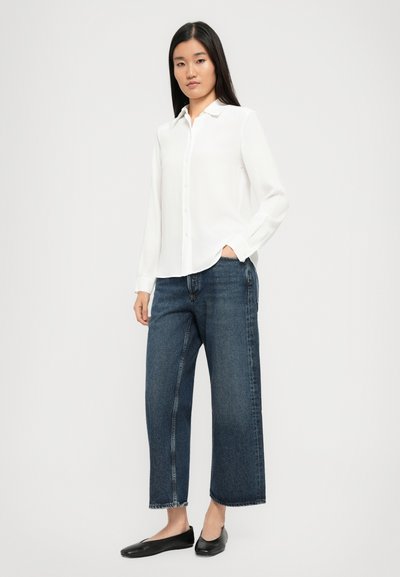 White, long-sleeve, button-up shirt with a collar, paired with loose, dark blue denim jeans and black flat shoes. Simple and modern design.