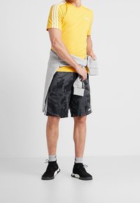 Camiseta atlética amarilla con tres franjas blancas, sudadera gris atada a la cintura, pantalones cortos de camuflaje negros y zapatillas negras.