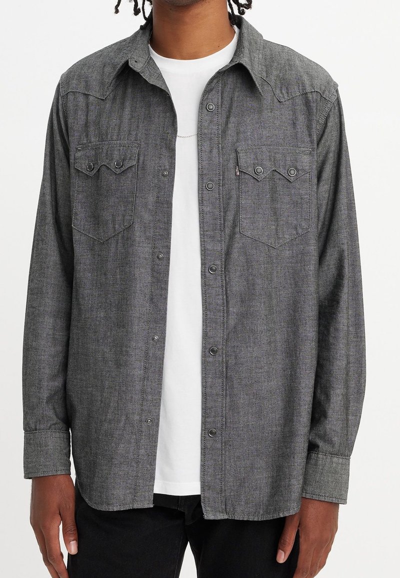 Homme portant une chemise gris foncé à boutons par-dessus un t-shirt blanc avec deux poches poitrine et un pantalon noir.