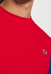 Mans nedre ansikte och hals bär en enkel röd t-shirt med en liten grön krokodillogo på bröstet.