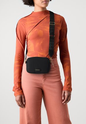 Vrouw draagt een roestkleurige top met lange mouwen en roze broek, met een zwarte rechthoekige crossbodytas die een brede, patroonrijke band over de borst heeft.