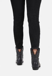 Bottines noires avec des détails cloutés, des orteils pointus et des côtés découpés. Portées avec un jean noir ajusté, mettant en valeur un matériau texturé.