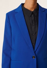 Modrý blazer s přiléhavým střihem, se zářezovými klopami a jedním knoflíkem. Nosený přes černou košili s límečkem a detaily na knoflících.