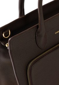 Sac à main en cuir marron foncé avec des poignées arrondies, surface texturée, poche zippée dorée et petit détail en anneau en métal doré.