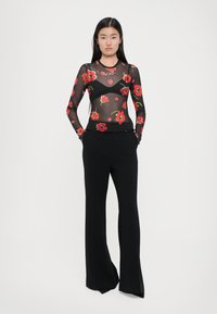 Sheer black long-sleeve top with red floral print and scalloped hem, paired with wide-leg black trousers. Model stands upright with hands in pockets.