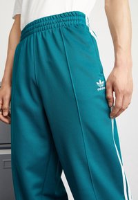 Turquoise joggings met een gestructureerde afwerking, elastische tailleband, witte zijkanten en een klein wit Adidas-logo op de linker dij.
