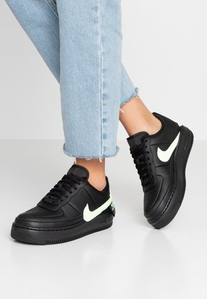Osoba ubrana w jasnoniebieskie dżinsy i czarne buty Nike z podświetlanymi białymi logo Swoosh, stojąca na szarej podłodze.