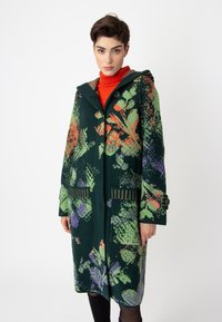 Hooded coat in dark green with floral pattern in orange, purple, and green. Features large pockets and button detailing on sleeves.