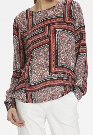 Femme portant un haut à manches longues avec des motifs géométriques et cachemire rouges, noirs et blancs, associé à un pantalon blanc.