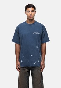 T-shirt in cotone blu oversize con scollatura rotonda, dettagli scoloriti e logo stampato sul petto. Abbinato a pantaloni in denim scuro.