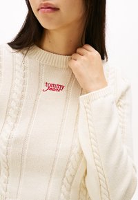 Kremowy sweter z dzianiny z wzorami ściągaczowymi i warkoczowymi, z małym czerwonym logiem "tommy jeans" przy dekolcie oraz wysokim kołnierzem.