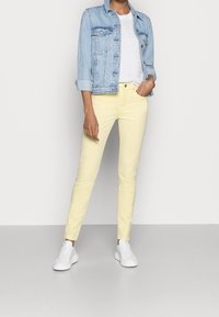 Jeans ajustados de color amarillo claro combinados con una chaqueta de mezclilla azul clara y una camiseta blanca, completados con zapatillas blancas.