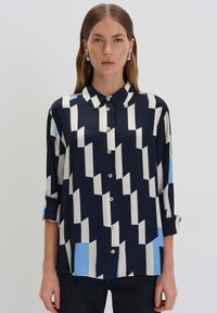 Camicia blu navy con motivi geometrici bianchi e azzurri, maniche lunghe, colletto classico e chiusura con bottoni. Tessuto morbido e leggero.