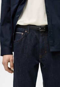 Mørke denimjeans med lige snit, der har kontrastsyninger, et sort bælte og en marineblå blazer over en lys skjorte.