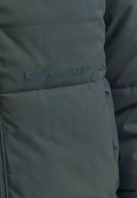 Tamnozelena puffer jakna glatke teksture. Sadrži suptilan logo "Originals Studio" ugraviran na površini. Minimalistički dizajn.