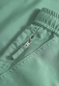 Zöld szövet közeli kép, cipzáros zsebbel, sima textúrával és elasztikus derékrész részletekkel. Tiszta varrás és modern kialakítás.