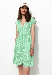 Robe florale verte à manches courtes, décolleté en V, taille cintrée et boutons sur le devant. Portée avec une casquette blanche et un bracelet en argent. Fond uni.