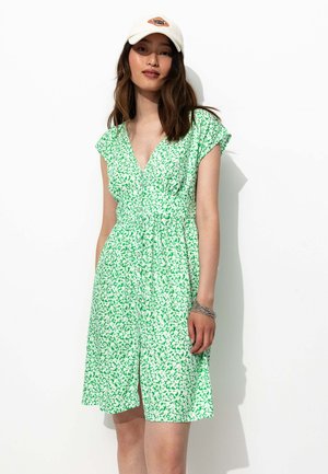 Robe florale verte à manches courtes, décolleté en V, taille cintrée et boutons sur le devant. Portée avec une casquette blanche et un bracelet en argent. Fond uni.
