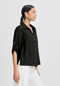 Camicia nera con colletto e maniche a tre quarti, realizzata in un tessuto texturizzato e con spacchi laterali, abbinata a pantaloni bianchi.