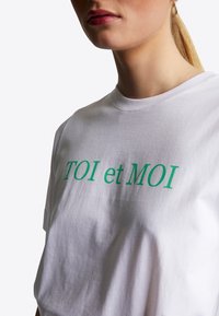 Vit bomulls t-shirt med grön text "TOI et MOI." Klassisk rundhalsad design med korta ärmar. Slen yta och avslappnad passform.