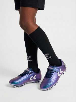 Jambes portant des chaussettes noires montantes et des crampons de football Hummel violets avec des logos chevrons blancs sur fond blanc.