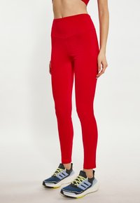 Rote hohe Leggings aus dehnbarem Stoff mit nahtlosem Design, kombiniert mit blau-schwarzen Sportschuhen mit gelben Akzenten.