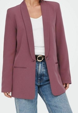 Femme portant un blazer mauve sur un haut blanc avec un jean bleu et une ceinture noire à boucle dorée.